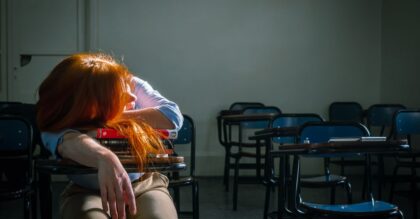 A tired student with red hair sleeps in an empty classroom under soft natural light.