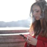 Portrait of a woman in red coat smoking and texting outside on a sunny day.