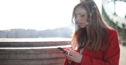 Portrait of a woman in red coat smoking and texting outside on a sunny day.