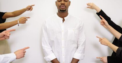 A calm man in white stands with closed eyes while several hands point at him in concept of accusation.