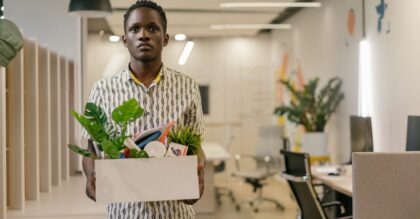 A serious man carries a box of belongings in an office setting, symbolizing dismissal or job change.